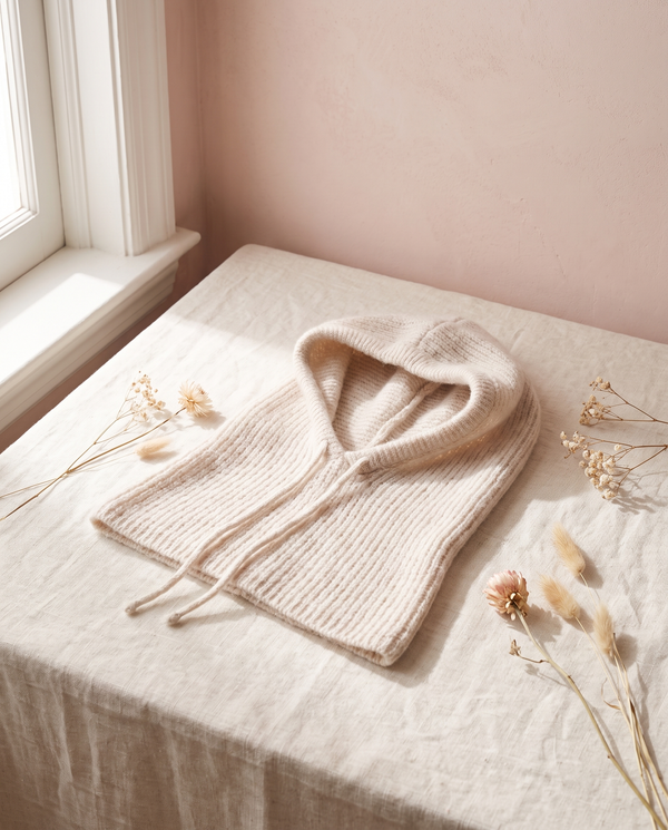 Cagoule maille Primerose écru, posée sur un lin crème, mur rose poudré